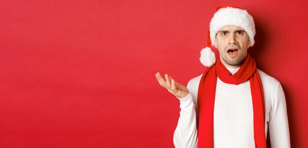 Concept of christmas winter holidays and celebration Portrait of confused man in santa hat and scarf frowning and looking perplexed standing over red background