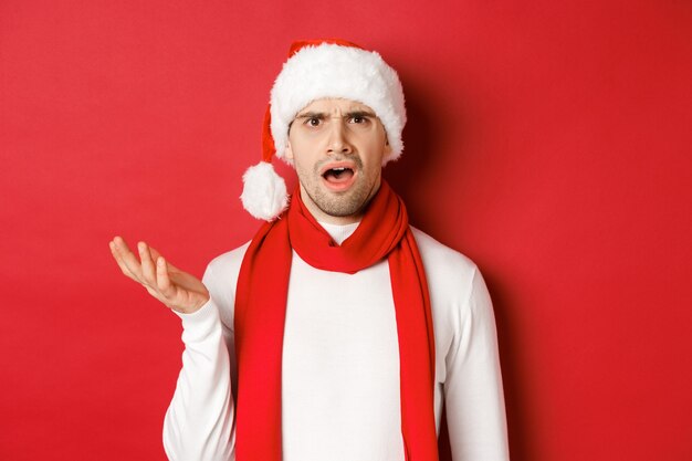 크리스마스, 겨울 방학 및 축하의 개념입니다. 산타 모자와 스카프를 쓴 혼란스러운 남자의 초상화, 인상을 찡그린 채 당황한 표정으로 빨간색 배경 위에 서 있습니다.