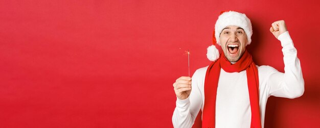 Concept of christmas winter holidays and celebration Portrait of excited handsome man raising hand up and holding sparkler wishing happy new year standing over red background