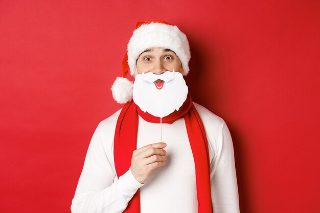Концепция Рождества, зимних праздников и празднования. Портрет забавного человека в шляпе санта-клауса, держащего маску бороды, наслаждающегося новогодней вечеринкой, стоящего на красном фоне