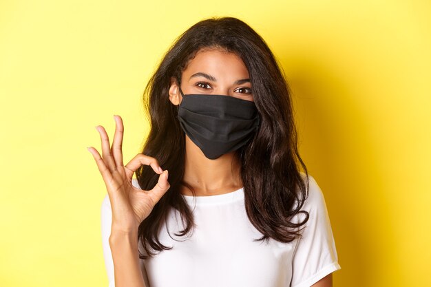 Понятие о коронавирусе, пандемии и образе жизни. Крупный план довольно афро-американской женщины в черной маске для лица, показывая хорошо, войдите в одобрение, хвалите хорошую работу, желтый фон.