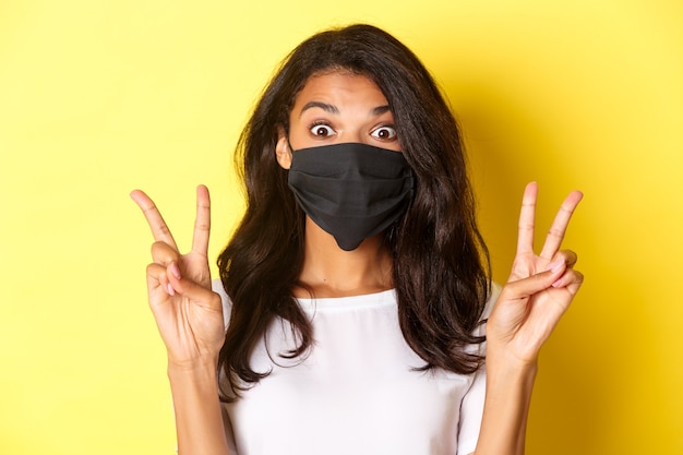 Free photo concept of coronavirus, pandemic and lifestyle. close-up of silly african-ameircan girl in black face mask, showing peace signs and standing over yellow background