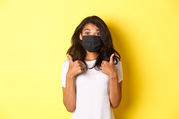 Concept of coronavirus, pandemic and lifestyle. Portrait of beautiful african-american girl in black face mask, showing thumbs-up in approval, like something good, yellow background.