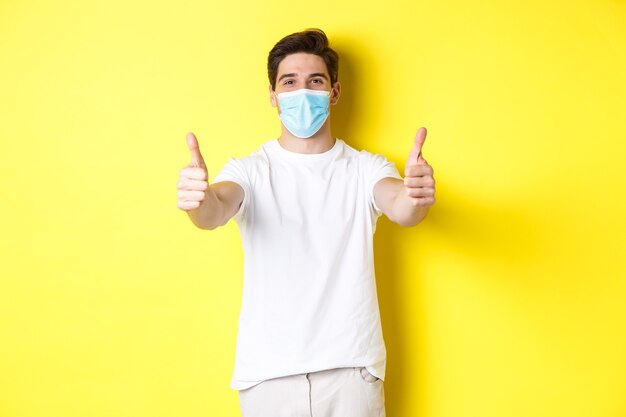 Concept of coronavirus, pandemic and social distancing. Confident man protecting himself from covid-19 with medical mask, showing thumbs up in approval, yellow background