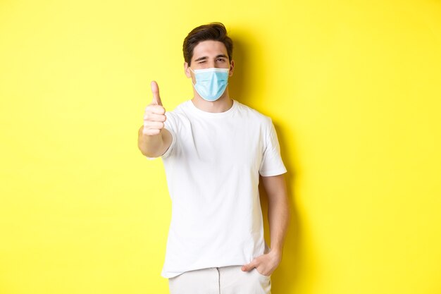 Concept of coronavirus, pandemic and social distancing. Confident young man in medical mask showing thumbs up and winking, yellow background.