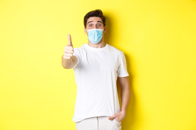 Концепция коронавируса, пандемии и социального дистанцирования. Под впечатлением парень в медицинской маске показывает большой палец вверх в знак одобрения, как будто что-то потрясающее, желтый фон.