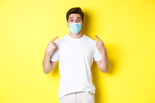 코로나바이러스, 전염병 및 사회적 거리의 개념. 얼굴에 의료 마스크를 가리키는 젊은 놀란 남자, 노란색 배경