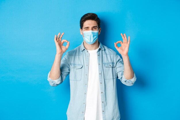 コロナウイルス、検疫および社会的距離の概念。医療用マスクのウィンクをしている生意気な男は、大丈夫な兆候を示し、何かを保証または保証します。