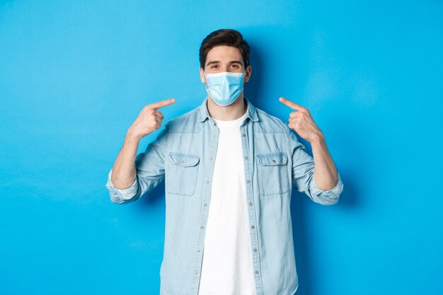 Concept of coronavirus, quarantine and social distancing. Handsome man pointing at medical mask and smiling, protection from virus spread during pandemic, blue background.