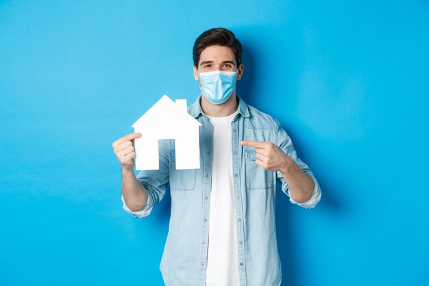 Concept of coronavirus, quarantine and social distancing. Young man searching aparment for rent, business loans, pointing at house model, wearing medical mask, blue background