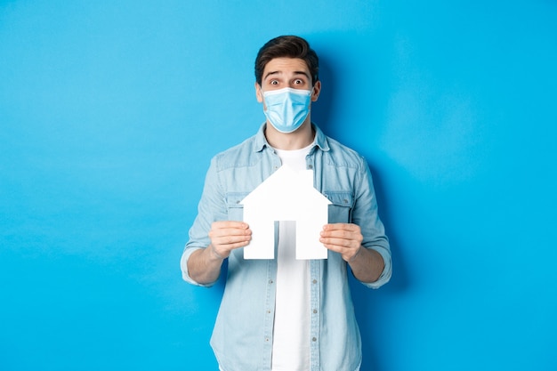 Concept of coronavirus, quarantine and social distancing. Young man searching apartment, showing house paper model, wearing medical mask, renting or buying propery, blue background