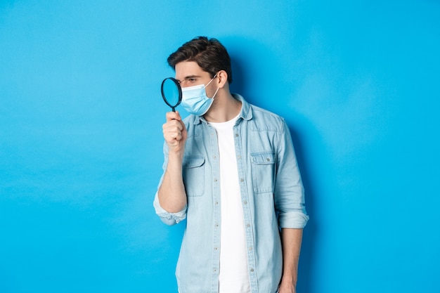 Concept of coronavirus, social distancing and pandemic. Man in medical mask searching for something, looking left through magnifying glass, studying copy space, blue background