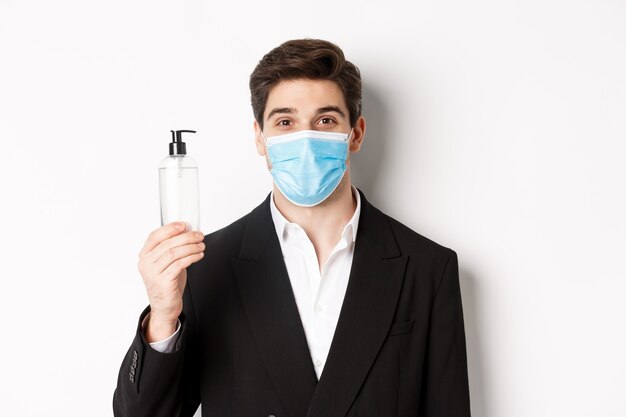 Concept of covid-19, business and social distancing. Close-up of handsome man in trendy suit and medical mask, showing hand sanitizer, standing against white background