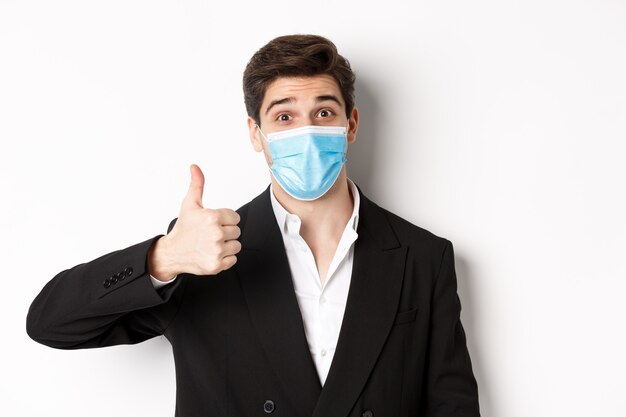Concept of covid-19, business and social distancing. Close-up of happy businessman in black suit and medical mask, showing thumbs-up, making a compliment, white background