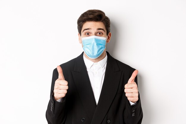 Concept of covid-19, business and social distancing. Close-up of happy businessman in black suit and medical mask, showing thumbs-up, making a compliment, white background