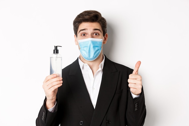 Concept of covid-19, business and social distancing. Close-up of satisfied handsome man in suit and medical mask, showing thumb-up and hand sanitizer, standing against white background.