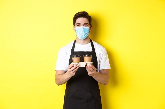 Concept of covid-19, cafe and social distancing. Barista in medical mask serving coffee in takeaway cups, standing in black apron against yellow background.