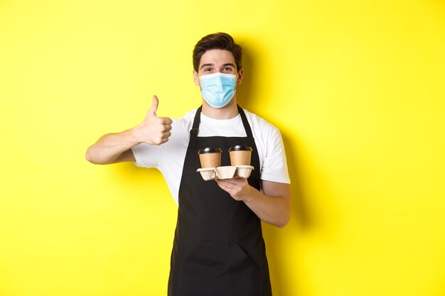 Concept of covid-19, cafe and social distancing. Young male barista in medical mask and black apron holding takeaway coffee cups, showing thumb up, yellow background.