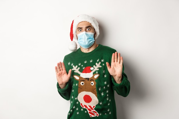 Concept of covid-19 and Christmas holidays. Anxious and freak out guy in santa hat with medical mask rejecting something, declining offer, standing over white background.