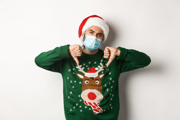 Concept of covid-19 and Christmas holidays. Displeased guy in face mask and santa hat showing thumbs down, standing over white background