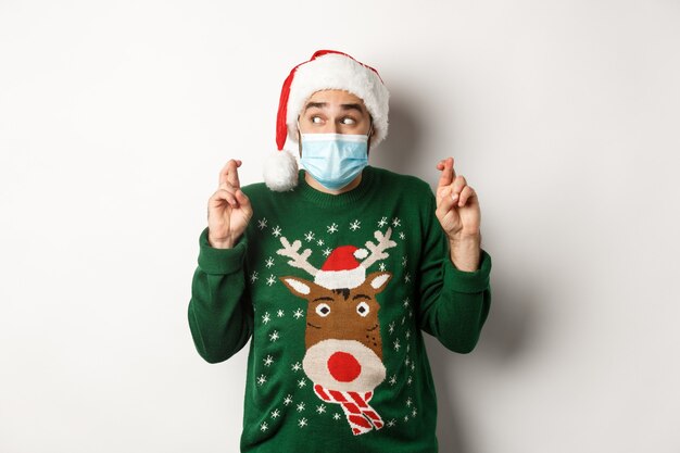 Concept of covid-19 and Christmas holidays. Excited man in face mask and Santa hat cross fingers, making a wish, standing over white background