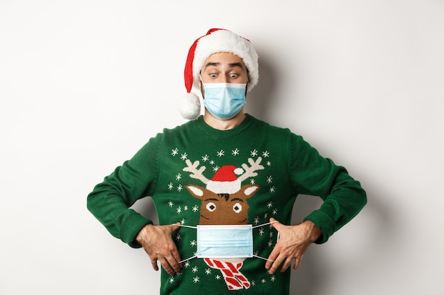 Concept of covid-19 and Christmas holidays. Funny man put on face mask on his sweater deer, standing over white background.