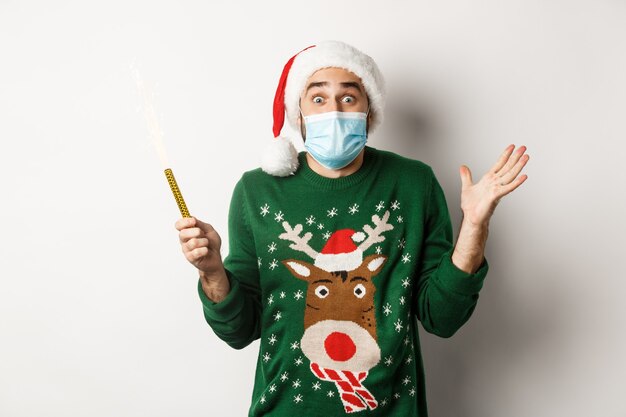 Concept of covid-19 and Christmas holidays. Happy young man in face mask celebrating Christmas, holding party sparkler and rejoicing, standing over white background