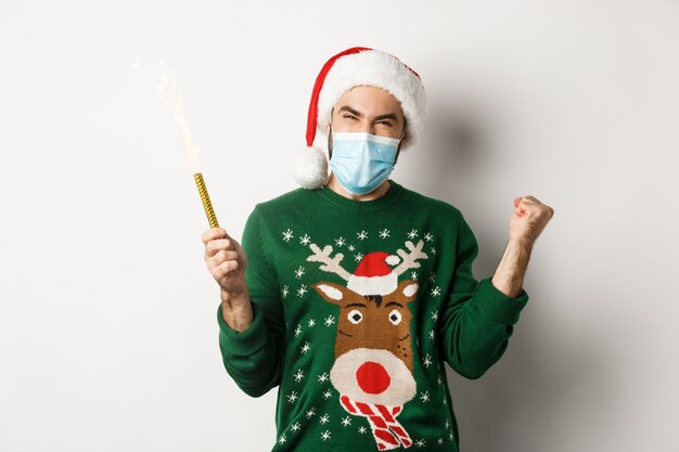 Concept of covid-19 and Christmas holidays. Happy young man in face mask celebrating Christmas, holding party sparkler and rejoicing, standing over white background.