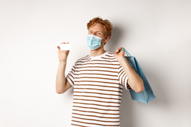 Concept of covid-19 and lifestyle. Happy young shopper in face mask holding shopping bag and showing plastic credit card, buying with discounts, white background