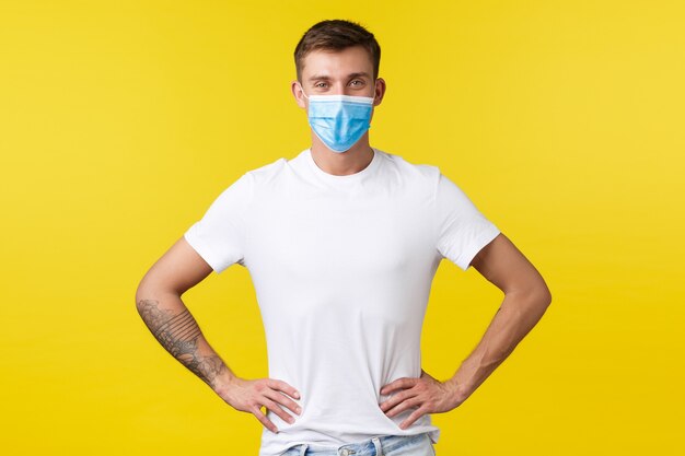 Concept of covid-19 pandemic outbreak, lifestyle during coronavirus social distancing. Confident handsome slim guy in medical mask and basic white t-shirt ready for work, hold hands on waist.