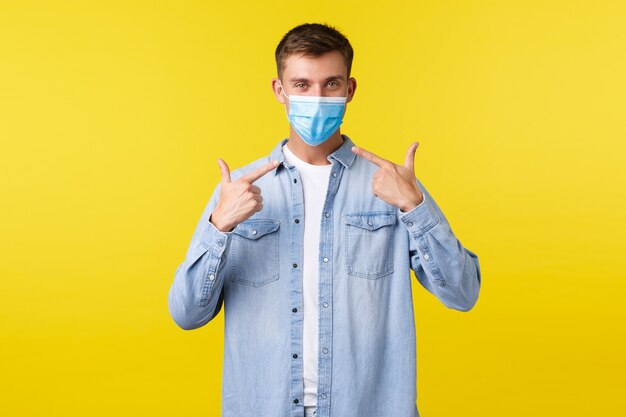 Concept of covid-19 pandemic outbreak, lifestyle during coronavirus social distancing. Smiling handsome man pointing at medical mask, recommend wearing it to prevent catching virus.