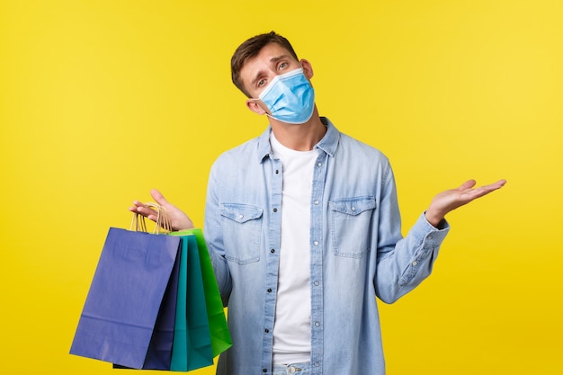 Free photo concept of covid-19 pandemic outbreak, shopping and lifestyle during coronavirus. indecisive, puzzled handsome guy in medical mask, shrugging clueless and carry bags from shop.