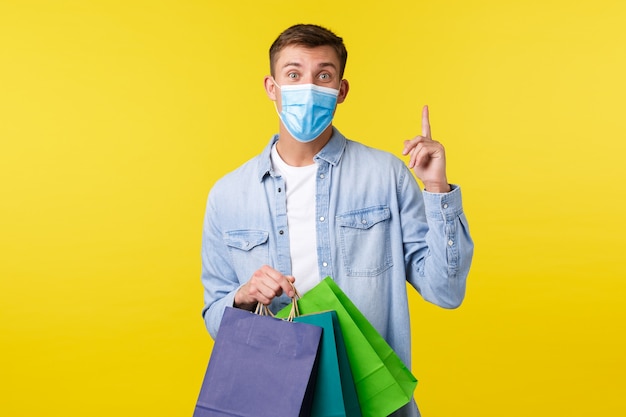Concept of covid-19 pandemic outbreak, shopping and lifestyle during coronavirus. young thoughtful man in medical mask, raising finger up, have suggestion or idea, holding bags from shop.
