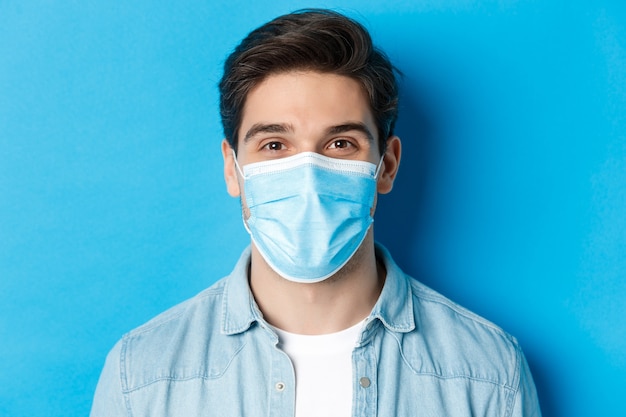 covid-19、パンデミックおよび検疫の概念。青い背景の上に立って、カメラを見ている医療マスクの幸せな男のクローズアップ。