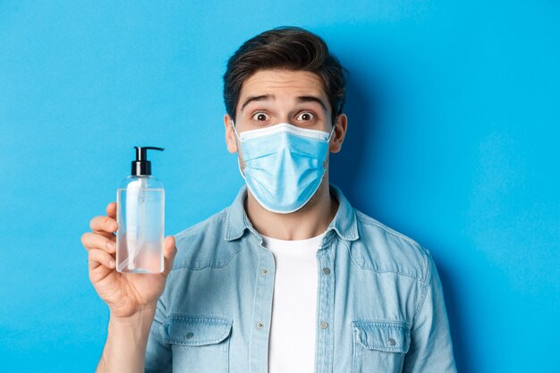 Концепция covid-19, пандемии и карантина. Удивленный парень в медицинской маске держит бутылку с дезинфицирующим средством для рук, изумленно приподнимая брови, стоя на синем фоне