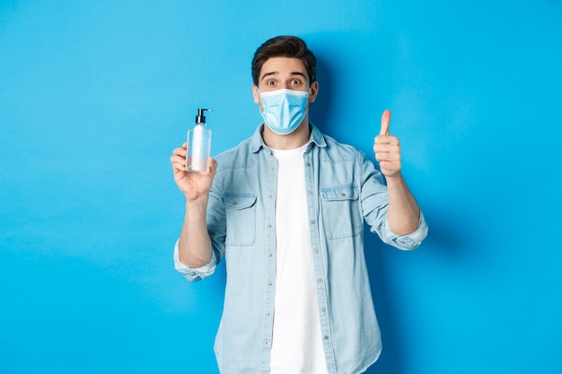 Concept of covid-19, pandemic and social distancing. Cheerful man in medical mask, showing hand sanitizer and thumbs-up, recommending sanitizer, standing against blue background