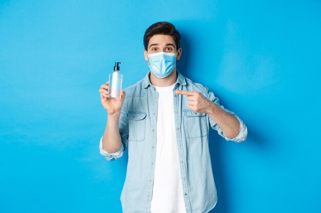 covid-19の概念、パンデミックおよび社会的距離。医療マスクのハンサムな男は、青い背景の上に立って、消毒剤を指して、手の消毒剤を使用するようにアドバイスします