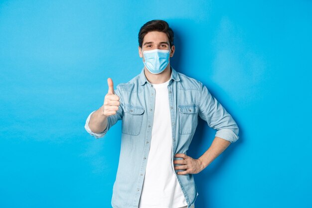 Concept of covid-19, pandemic and social distancing. Satisfied guy in medical mask showing thumb up in approval, standing against blue background