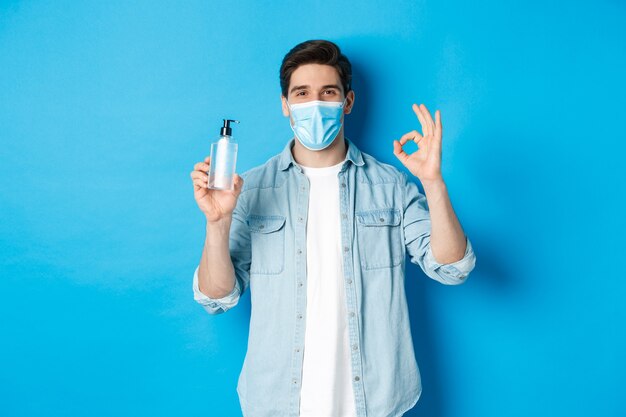 Concept of covid-19, pandemic and social distancing. Satisfied young man in medical mask recommending hand sanitizer, showing ok sign and antiseptic, standing against blue background