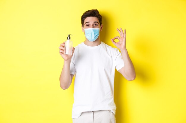 Concept of covid-19, quarantine and lifestyle. Satisfied young man in medical mask showing good hand sanitizer, make okay sign and recommending antiseptic, yellow background