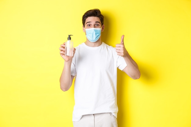 Concept of covid-19, quarantine and lifestyle. Satisfied young man in medical mask showing good hand sanitizer, thumbs up and recommending antiseptic, yellow background