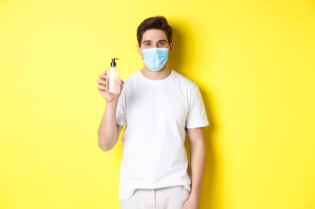 covid-19の概念、検疫およびライフスタイル。黄色の背景の上に立って、手の消毒剤、手の消毒製品を示す医療マスクの若い男。