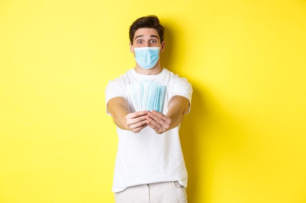 Concept of covid-19, quarantine and preventive measures. Young caucasian man giving medical masks for you, standing against yellow background.