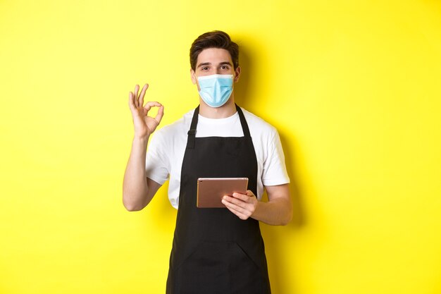 Концепция covid-19, малого бизнеса и пандемии. Продавец в медицинской маске и черном фартуке показывает знак ОК, принимает заказы с цифровым планшетом, желтым фоном.