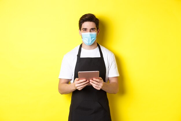 Концепция covid-19, малого бизнеса и пандемии. Официант в черном фартуке и медицинской маске принимает заказ, держа цифровой планшет, стоя на желтом фоне.