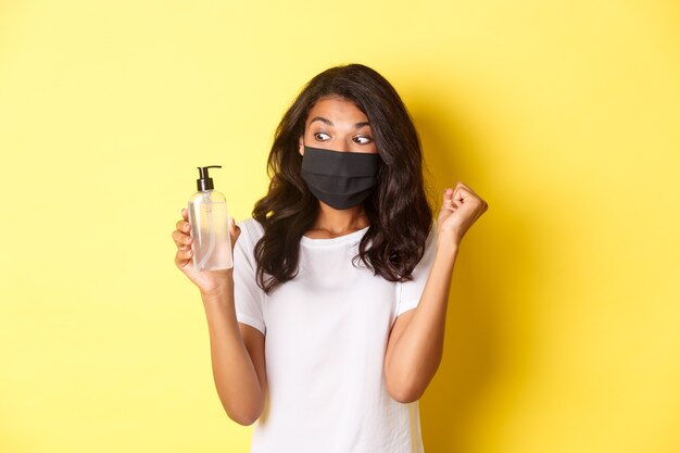 covid-19, 사회적 거리 및 생활 방식의 개념. 얼굴 마스크와 흰색 티셔츠를 입은 행복한 아프리카계 미국인 여성의 이미지, 좋은 손 소독제를 보여주고 주먹 펌프를 만드는 노란색 배경.