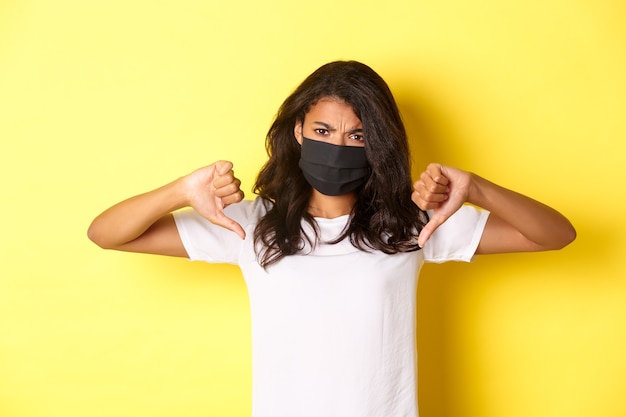 Концепция covid-19, социальное дистанцирование и образ жизни. Изображение расстроенной афро-американской протестующей женщины в черной маске, показывает палец вниз и разочарованно хмурится, желтый фон.