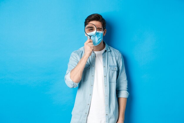 Concept of covid-19, social distancing and quarantine. Young man in medical mask searching for something, looking through magnifying glass, standing against blue background