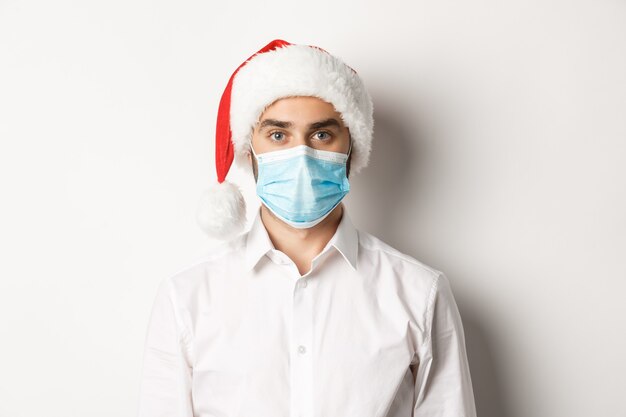Concept of covid-19, social distancing and winter holidays. Close-up of young man wearing santa hat and face mask from coronavirus, celebrating New Year 