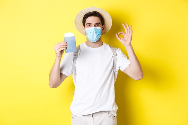 Concept of covid-19, tourism and pandemic. Happy male tourist in medical mask showing passport, going on vacation during coronavirus, make ok sign, yellow background